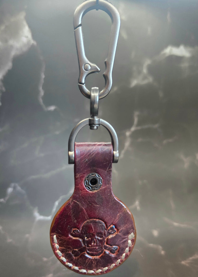Brown veg-tanned leather apple air tag case with a skull and crossbones stamped into it hanging on a solid stainless steel lobster clasp keychain.