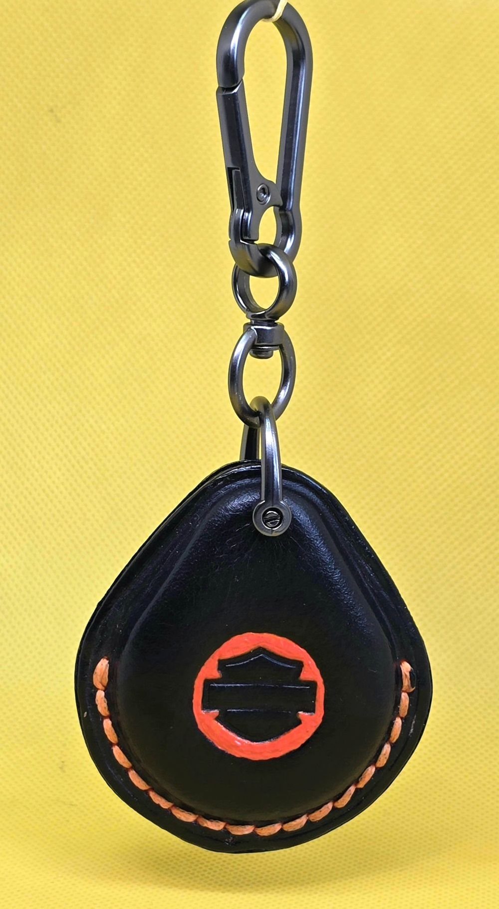 Black veg-tan leather key fob case for Harley Davidson motorcycles with orange stitching and logo hanging on a stainless steel lobster clasp keychain.