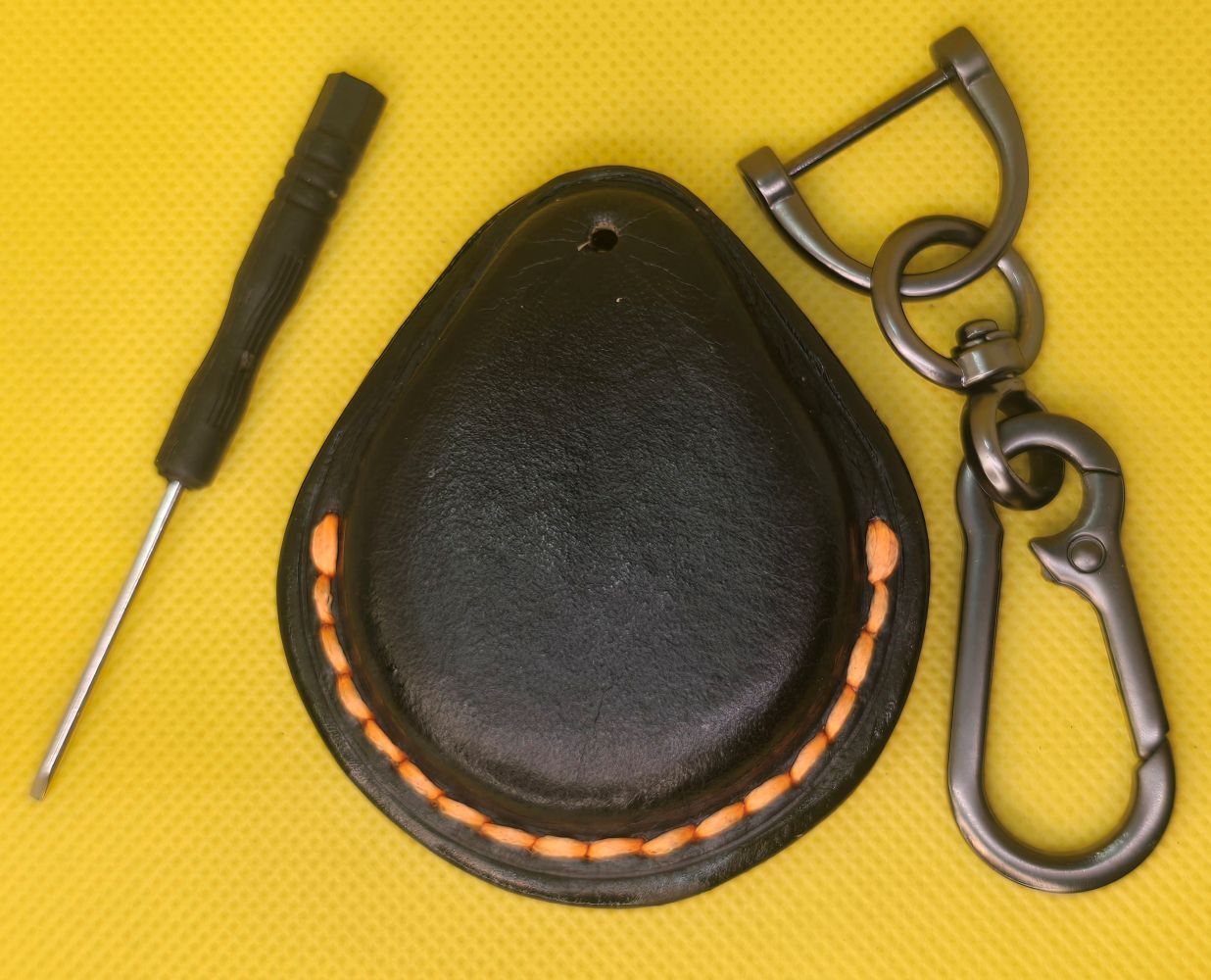 Back view of a black veg-tan leather key fob case for Harley Davidson motorcycles with orange stitching on a table with a lobster clasp keychain and mini screwdriver.