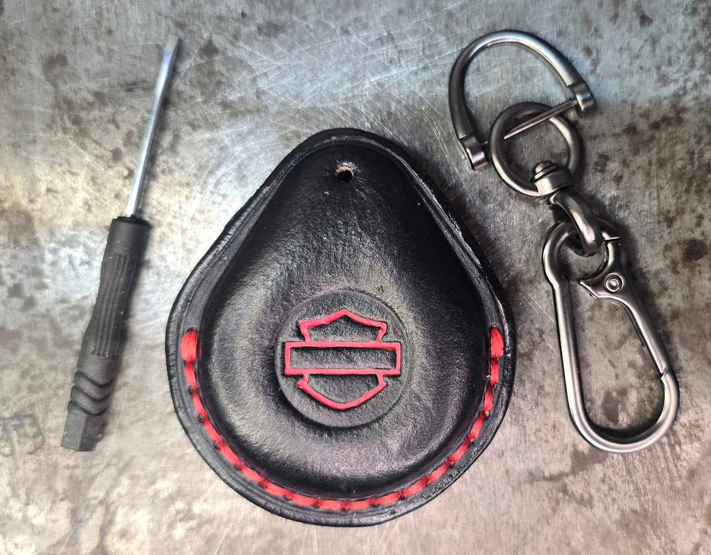 Picture of a black and red vegetable tanned leather key fob case for Harley Davidson motorcycles with red stitching and logo on a table with the included stainless steel keychain and screwdriver.