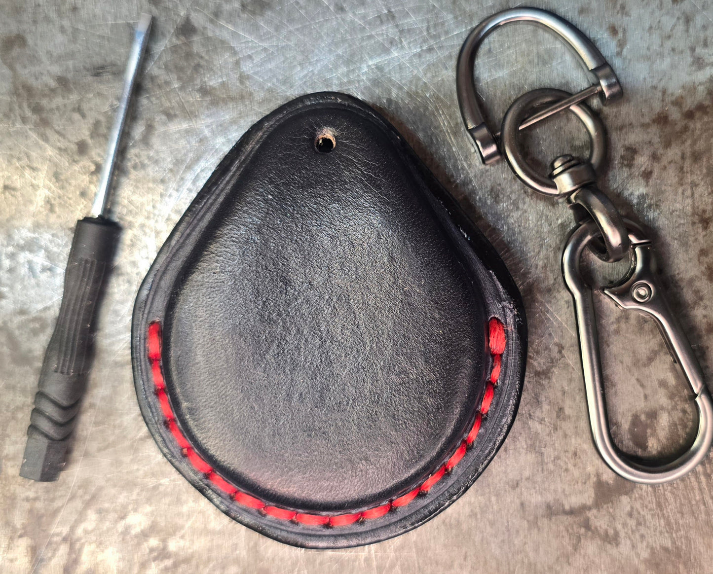 Back view of a black and red veg-tan leather key fob case for Harley Davidson motorcycles with red stitching on a table with a lobster clasp keychain and included mini screwdriver.