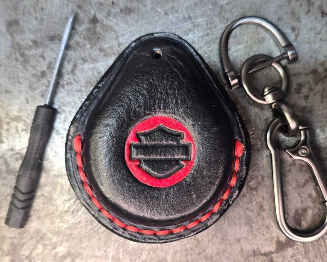 Picture of a black and red vegetable tanned leather key fob case for Harley Davidson motorcycles with red stitching on a table with the included stainless steel keychain and screwdriver.