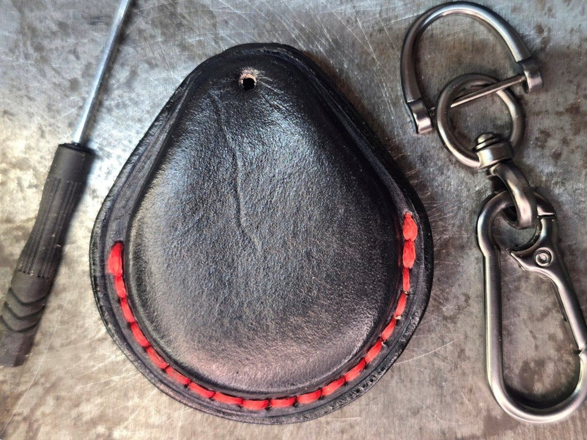 Back view of a black and red veg-tan leather key fob case for Harley Davidson motorcycles with red stitching on a table with a lobster clasp keychain and included mini screwdriver.