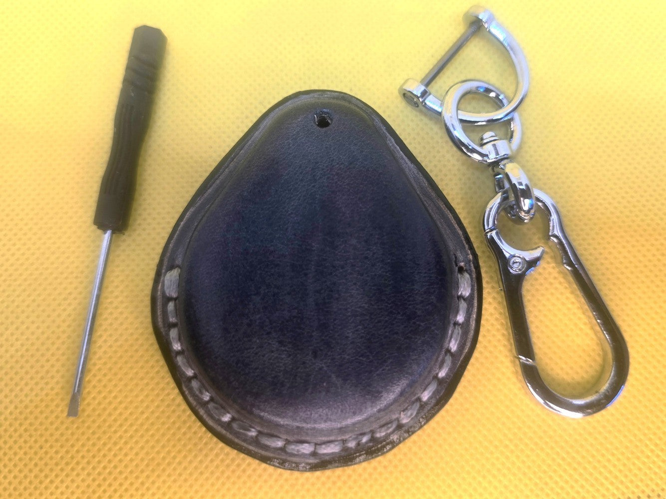 Back view of a gray veg-tan leather key fob case for Harley Davidson motorcycles with light gray stitching on a table with lobster clasp keychain and mini screwdriver.