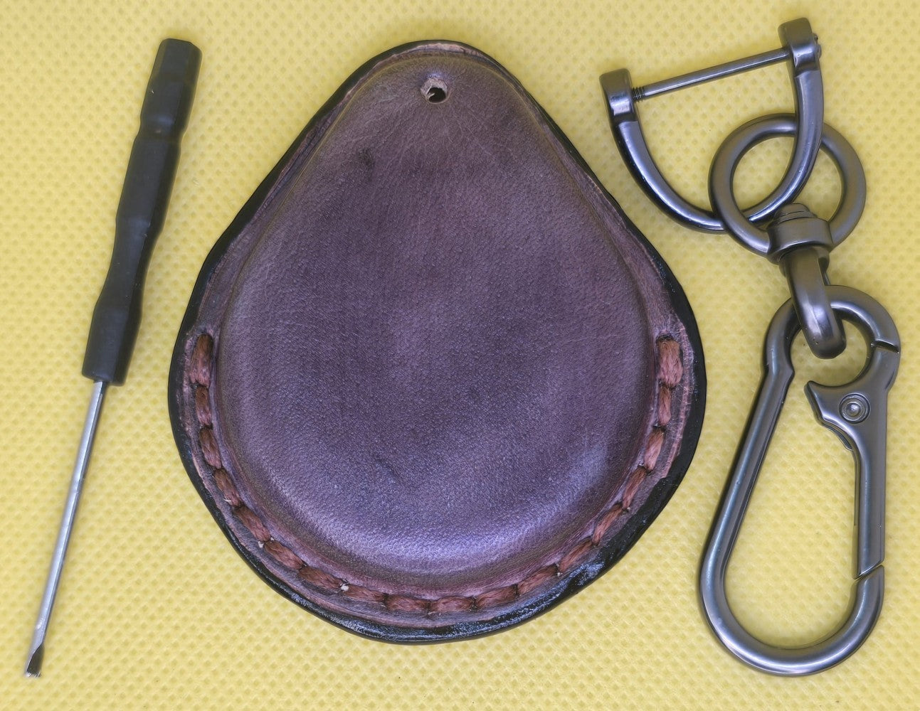 Back view of a mahogany veg-tan leather key fob case for Harley Davidson motorcycles on a table with a lobster clasp keychain and mini screwdriver.