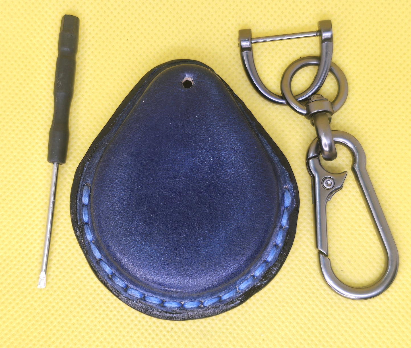 Back view of a navy blue veg-tan leather key fob case for Harley Davidson motorcycles with blue stitching on a table with a lobster clasp keychain and mini screwdriver.