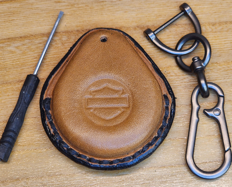 Light brown vegetable tanned leather key fob cases for Harley Davidson motorcycles with black stitching beside the included solid stainless steel lobster clasp keychain and small screwdriver.