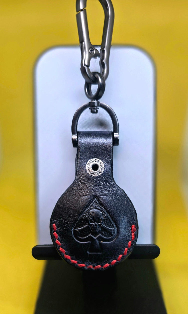 Black full-grain leather Apple airtag case with red stitching and a spade with skull and crossbones stamped into it.