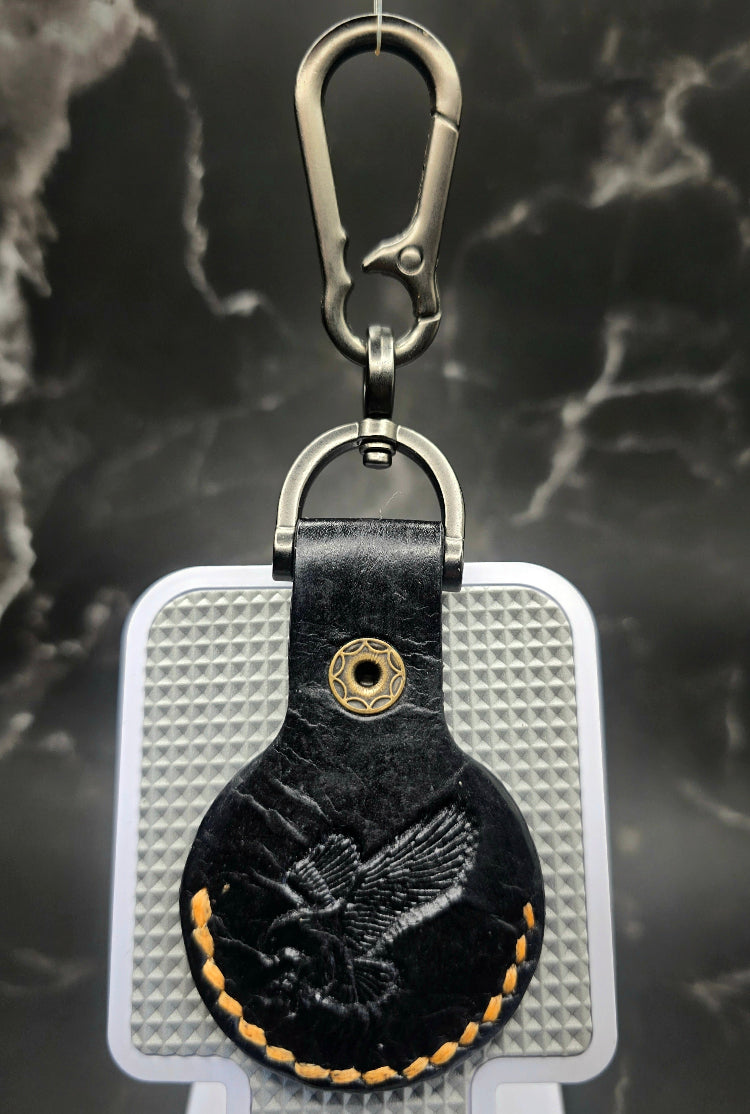 Black veg-tan leather apple airtag case with orange stitching and an eagle stamped on the back on a keychain.