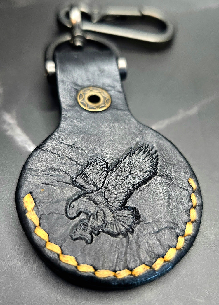 Black veg-tan leather apple airtag case with orange stitching and an eagle stamped on the back on a keychain.
