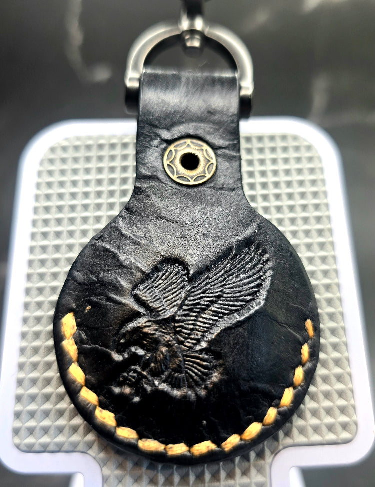 Black veg-tan leather apple airtag case with orange stitching and an eagle stamped on the back on a keychain.
