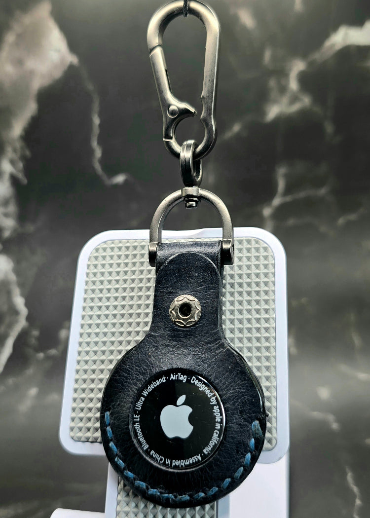 Black apple aie tag cover with blue stitching hanging on a lobster clasp keychain with air tag inserted into it.