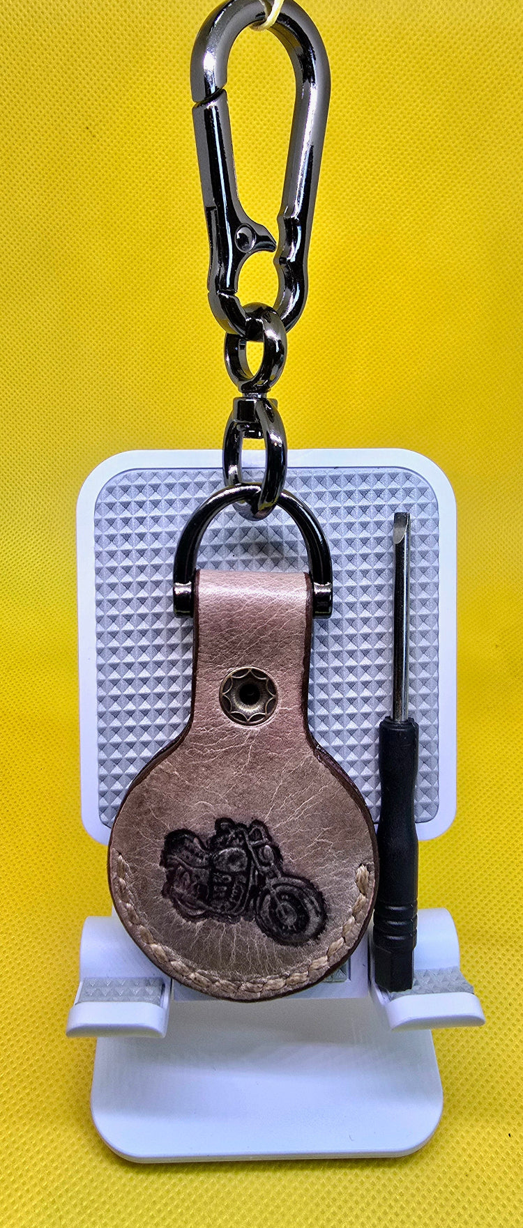 Brown Veg-tan leather apple airtag case with a motorcycle stamped into it on a stainless steel keychain with a small screwdriver next to it.