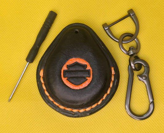 Picture of a black vegetable tanned leather key fob case for Harley Davidson motorcycles with orange logo and stitching on a table with the included stainless steel keychain and screwdriver.