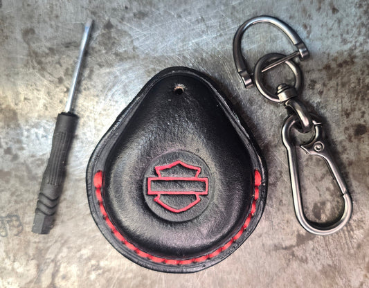 Picture of a black and red vegetable tanned leather key fob case for Harley Davidson motorcycles with red stitching and logo on a table with the included stainless steel keychain and screwdriver.