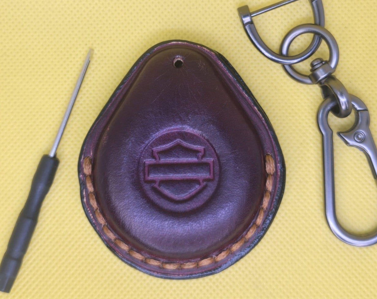 Picture of a burgundy vegetable tanned leather key fob case for Harley Davidson motorcycles on a table with the included stainless steel keychain and screwdriver.