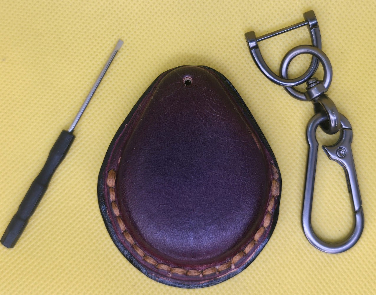 Back view of a burgundy veg-tan leather key fob case for Harley Davidson motorcycles on a table with a lobster clasp keychain and mini  screwdriver.