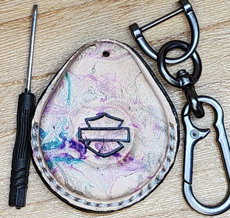 Front view of a premium vegetable tanned leather Harley Davidson key fob case with a unique tye dye look of purple and green with splashes of pink and edged in black. Solid stainless steel lobster clasp style keychain and mini screwdriver included in photo on each side.