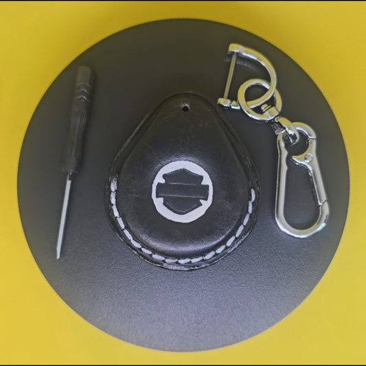 Video of a black and white veg-tan leather key fob case for Harley Davidson motorcycles with white stitching rotating on a display turntable with the included screwdriver and a stainless steel lobster clasp keychain next to it.