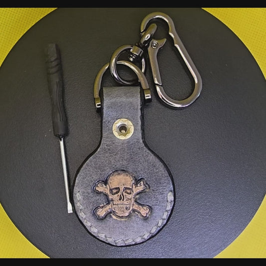 Video of a black colored vegetable tanned leather Apple air tag holder with a light brown skull and crossbones stamped into it on a lobster clasp keychain turning on a display pedestal with included screwdriver.