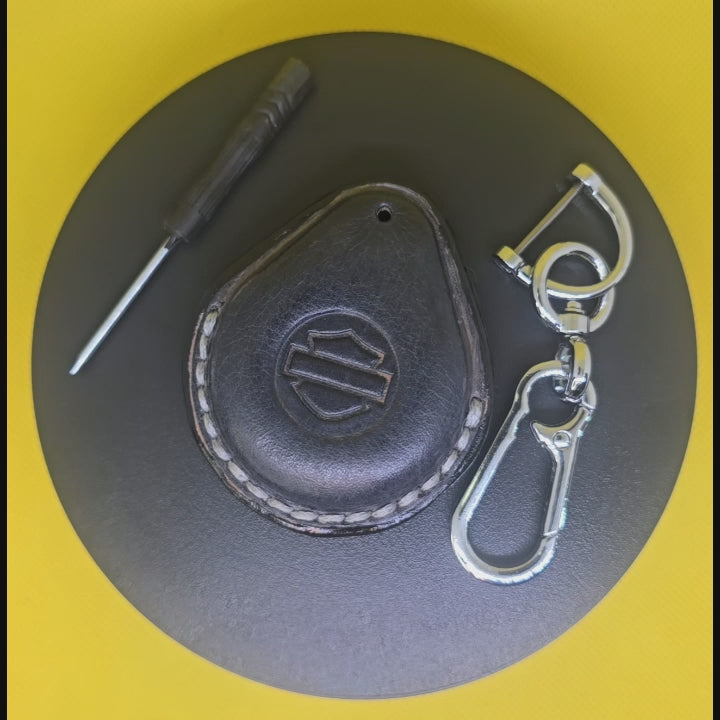 Video of a gray premium leather key fob cover for Harley Davidson motorcycles with light gray stitching rotating on a display turntable with a stainless steel lobster clasp keychain and screwdriver.