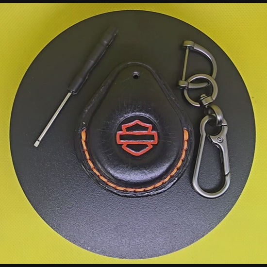 Video of a black Veg-tanned leather key fob case for Harley Davidson motorcycles with orange stitching and logo rotating on a turntable with the included solid stainless steel lobster clasp keychain and screwdriver.