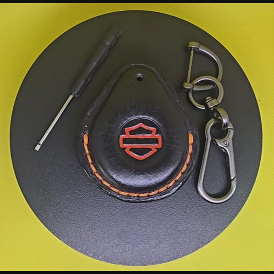 Video of a black Veg-tanned leather key fob case for Harley Davidson motorcycles with orange stitching and logo rotating on a turntable with the included solid stainless steel lobster clasp keychain and screwdriver.