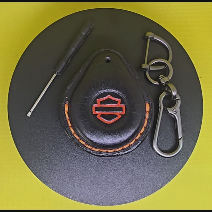 Video of a black Veg-tanned leather key fob case for Harley Davidson motorcycles with orange stitching and logo rotating on a turntable with the included solid stainless steel lobster clasp keychain and screwdriver.