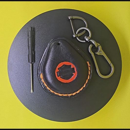Video of a black Veg-tanned leather key fob case for Harley Davidson motorcycles with orange logo and stitching rotating on a turntable with the included solid stainless steel lobster clasp keychain and screwdriver.