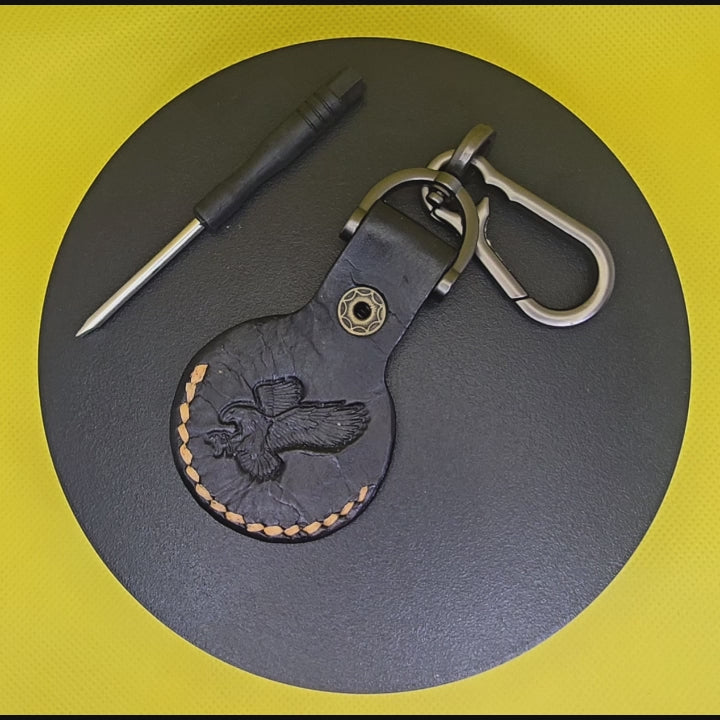 Black veg-tan leather apple airtag case with orange stitching and an eagle stamped on the back with a keychain rotating on a display turntable.