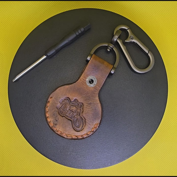 Video of a brown colored vegetable tanned leather Apple Airtag holder with a motorcycle stamped into it on a lobster clasp keychain turning on a display pedestal.