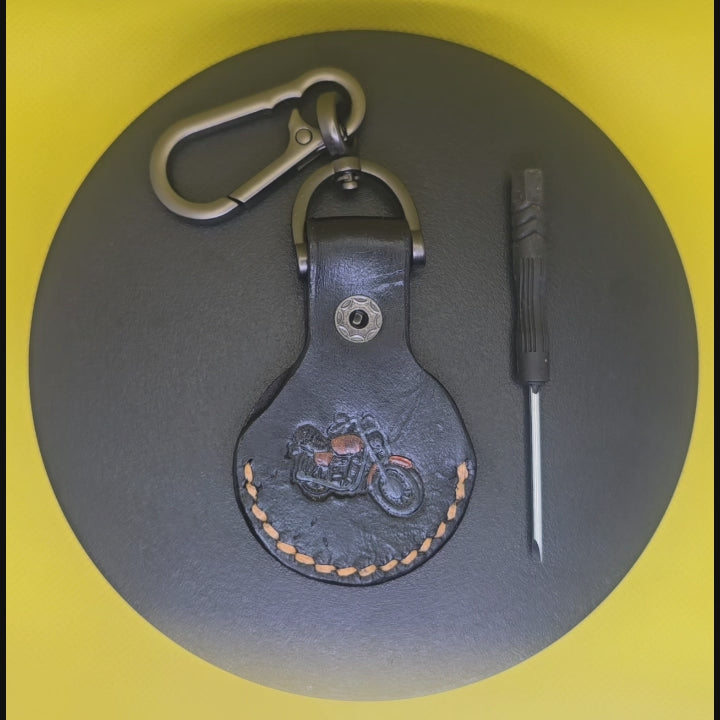 Video of black Veg-tan leather Apple AirTag case with keychain and orange stitching with black and orange motorcycle on a display turntable.