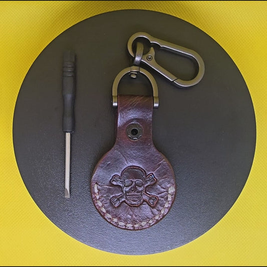 Video of a brown colored vegetable tanned leather Apple air tag holder with a skull and crossbones stamped into it on a lobster clasp keychain turning on a display pedestal with included screwdriver.