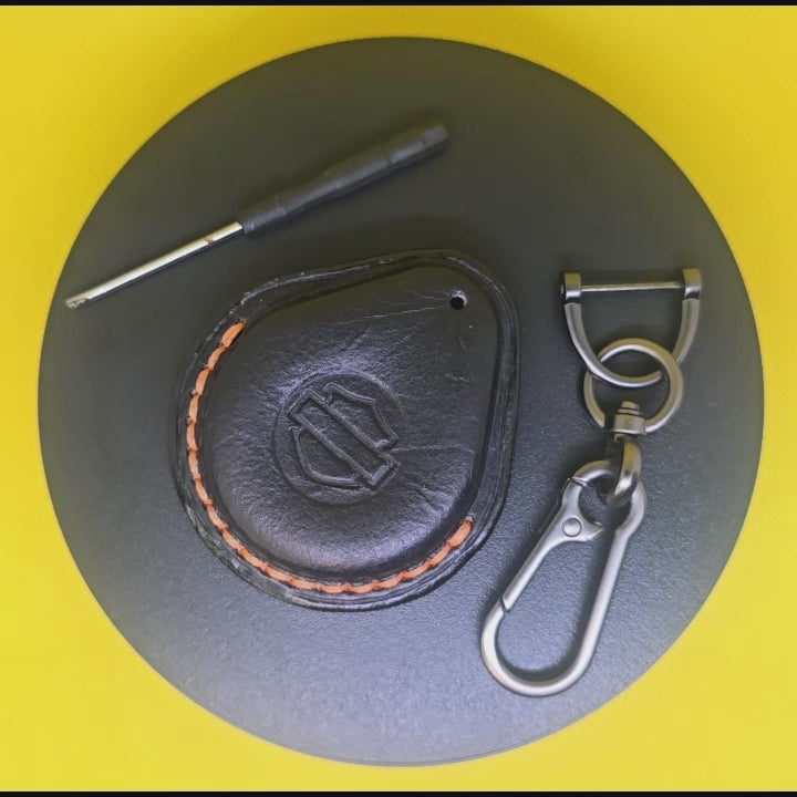 Video of a black Vegetable tanned leather key fob case for Harley Davidson motorcycles with orange stitching rotating on a pedestal with included stainless steel keychain and screwdriver.