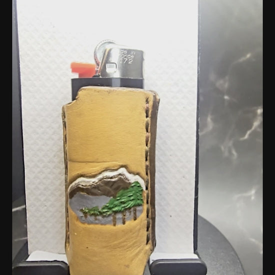Video of a veg-tan leather lighter case for bic lighters with a hand-painted mountain scene with trees.