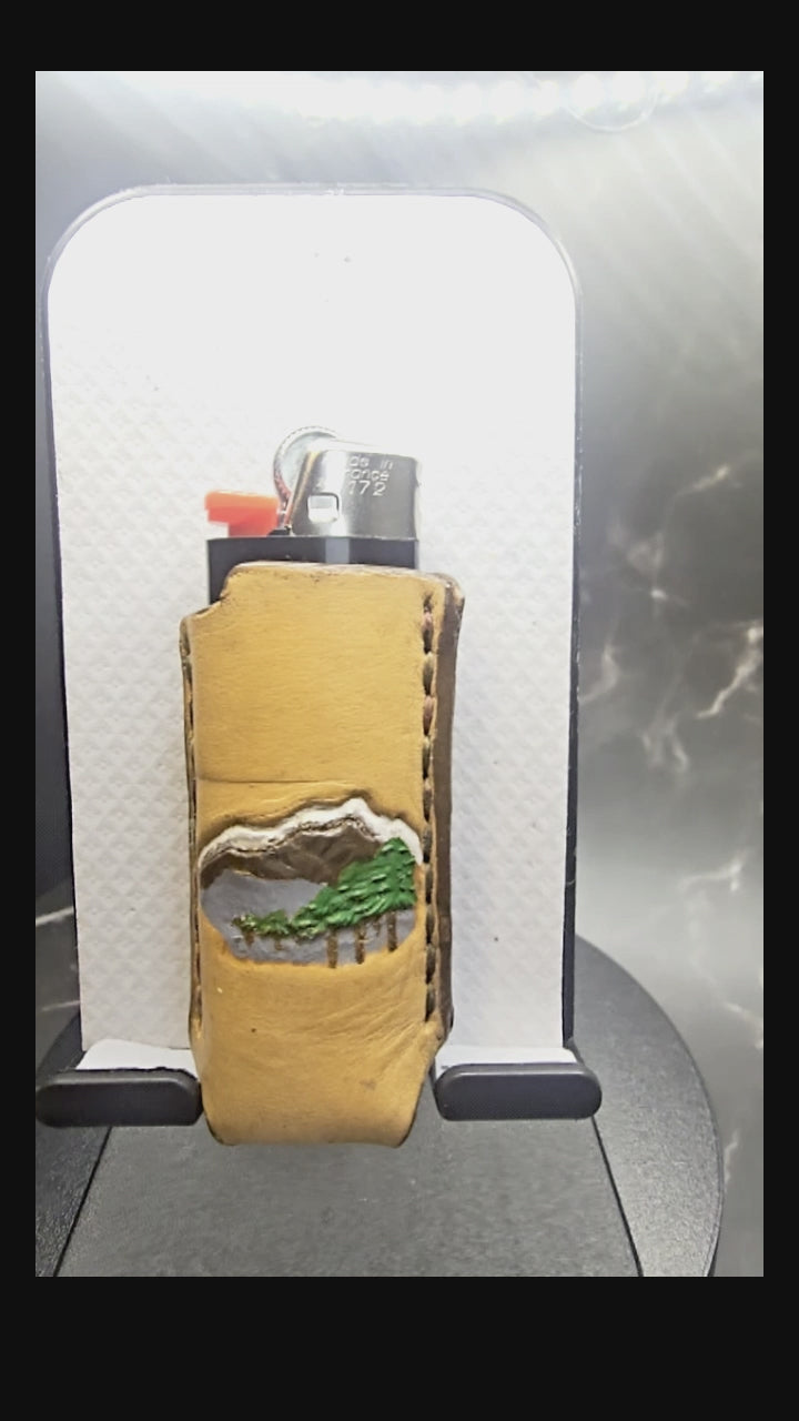 Video of a veg-tan leather lighter case for bic lighters with a hand-painted mountain scene with trees.