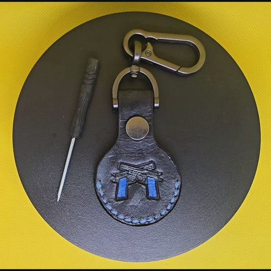 Video of a black colored vegetable tanned leather Apple Air tag holder with 2 guns with navy blue handles stamped into it on a lobster clasp keychain turning on a display pedestal with included screwdriver.