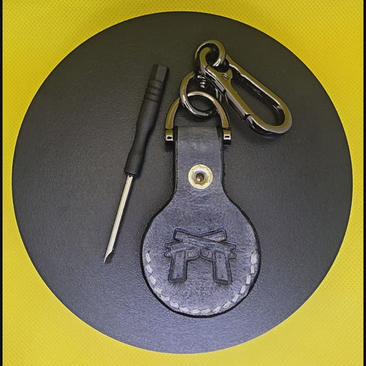 Video of a black colored vegetable tanned leather Apple Air tag holder with  2 guns stamped into it on a lobster clasp keychain turning on a display pedestal with included screwdriver.